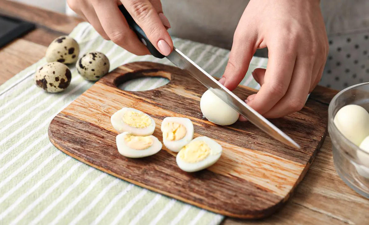 Hand slicing a peeled hard-boiled quail egg on a wooden cutting board, showing fully set yellow yolks and smooth whites, with speckled quail eggs in the background.
