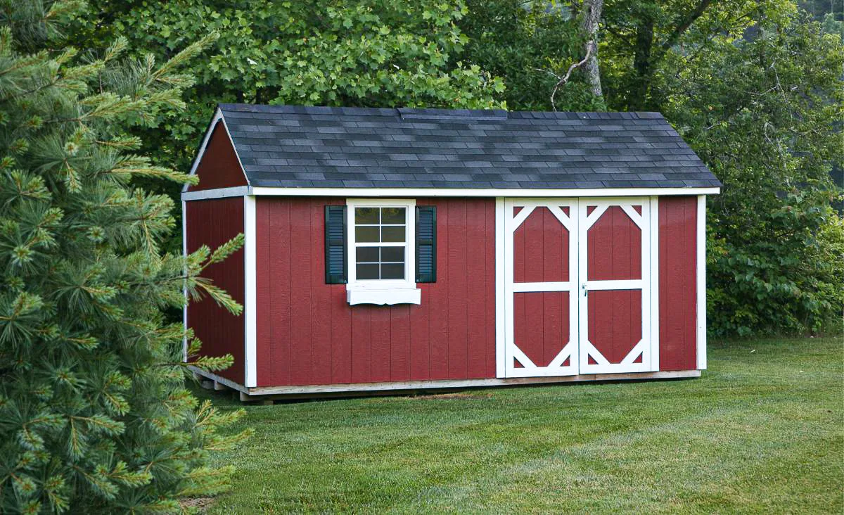 Red backyard storage shed with white trim, double barn-style doors, and a single window with black shutters, set on a grassy lawn and surrounded by trees, representing a potential standalone shed structure for housing Coturnix quail.