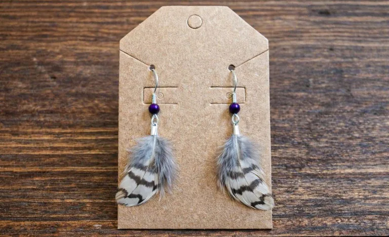 Handmade feather earrings displayed on a kraft paper jewelry card, featuring small gray and white barred quail feathers with deep purple beads, photographed against a rustic wooden tabletop.