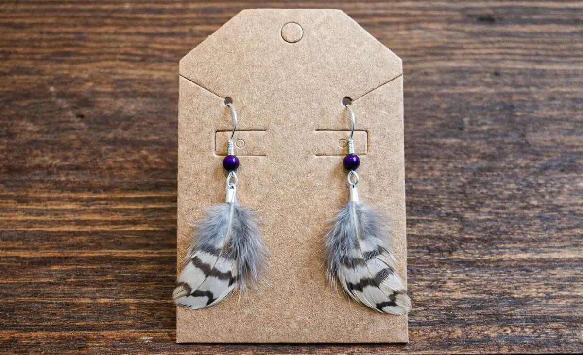 Handmade feather earrings displayed on a kraft paper jewelry card, featuring small gray and white barred quail feathers with deep purple beads, photographed against a rustic wooden tabletop.