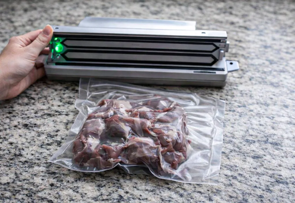 Vacuum sealing machine on a speckled granite countertop sealing a clear bag of raw quail meat, with a hand holding the machine in place and green indicator lights visible on the sealer.