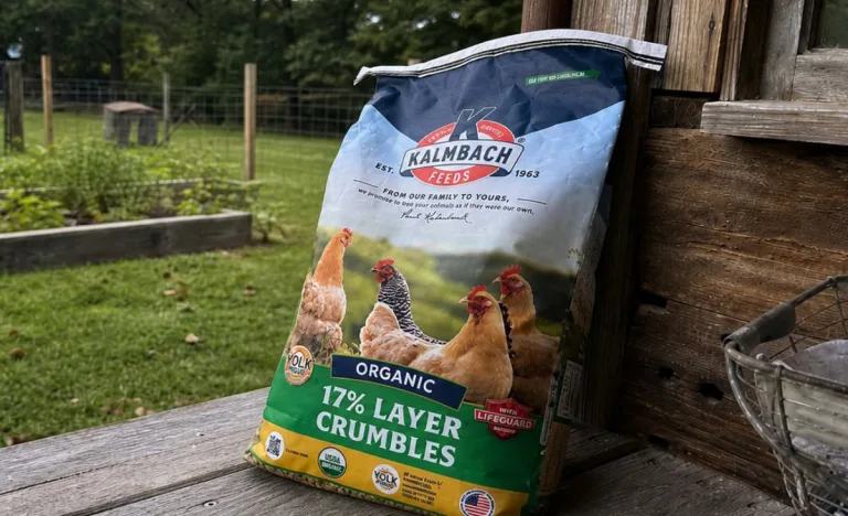 A bag of Kalmbach Organic 17% Layer Crumbles leaning against a rustic wooden wall on an outdoor homestead porch, with raised garden beds, fencing, and a grassy yard in the background.