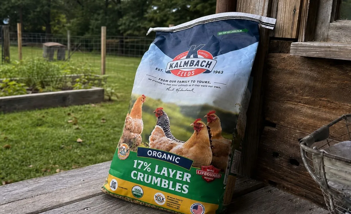 A bag of Kalmbach Organic 17% Layer Crumbles leaning against a rustic wooden wall on an outdoor homestead porch, with raised garden beds, fencing, and a grassy yard in the background.