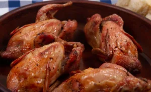 Four whole roasted Coturnix quail in a brown ceramic dish, browned with visible seasoning and pan juices, shown close up with bread in the background.
