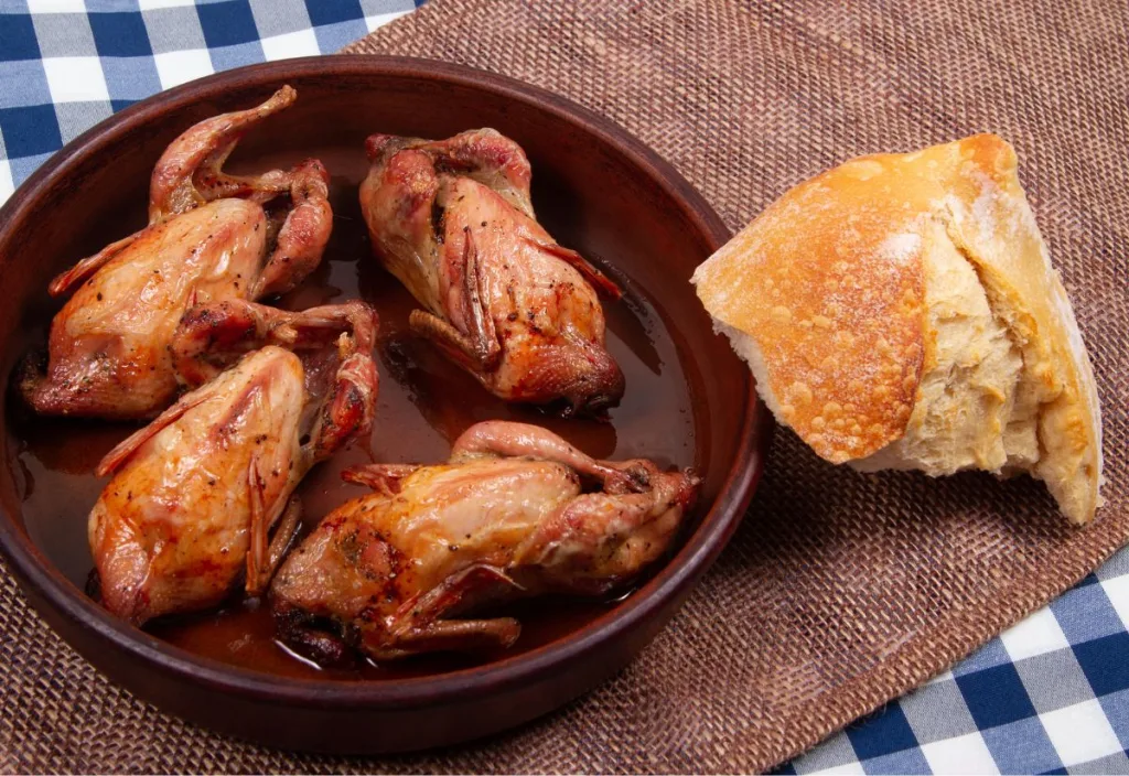 Whole roasted Coturnix quail served in a brown ceramic dish with pan juices, beside a piece of crusty bread on burlap and a blue plaid cloth.